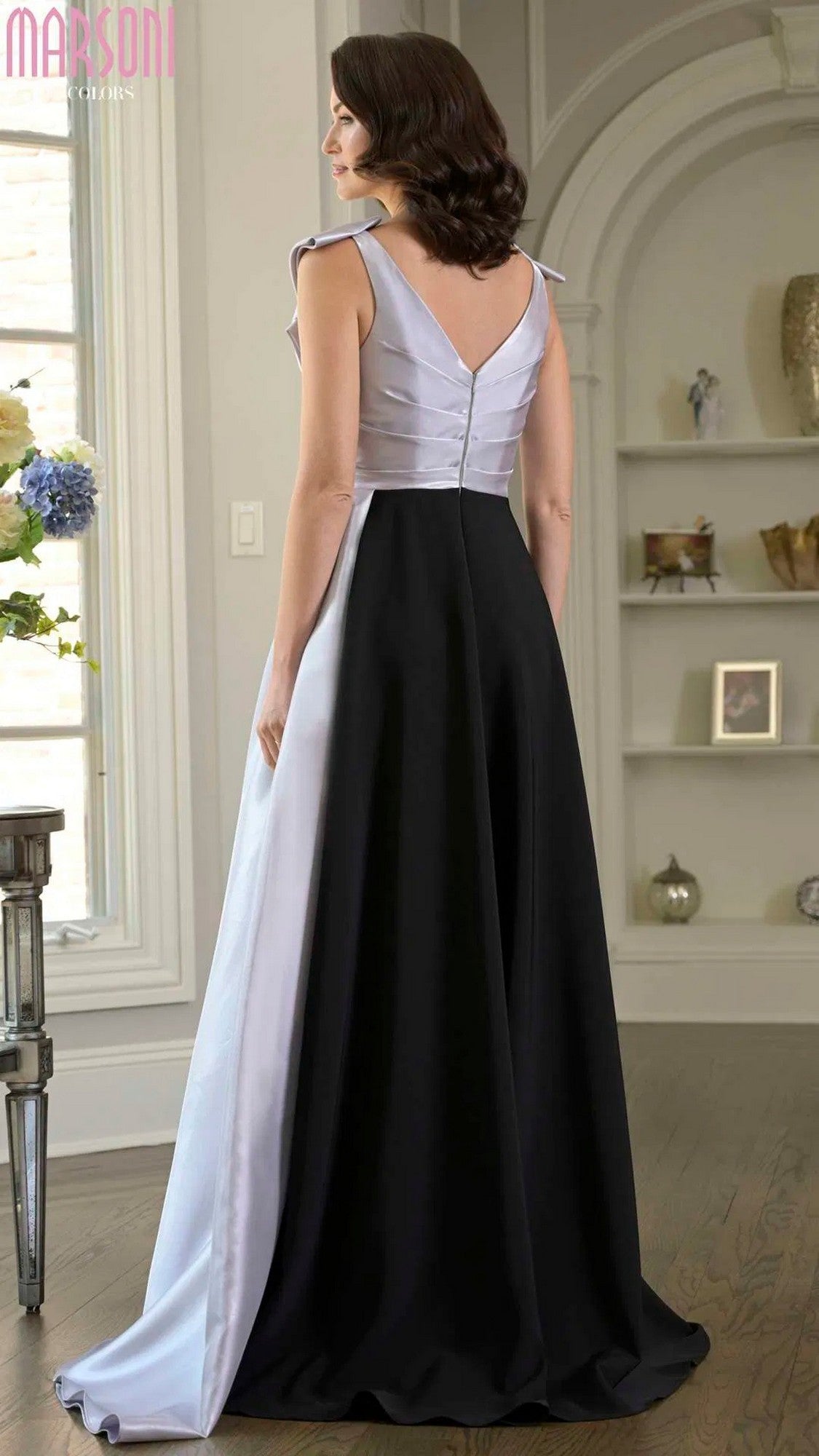 A woman stands indoors, viewed from the back, wearing the Marsoni M345 Long Formal Dress featuring a sleeveless, fitted white bodice and a long, flowing black A-line skirt.